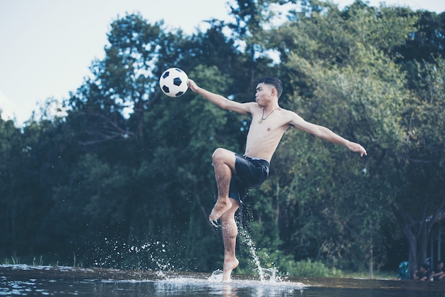 Rapaz asiático chuta uma bola de futebol em um rio