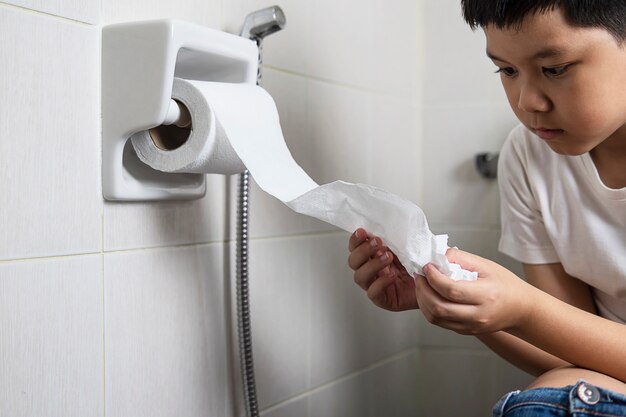 Rapaz asiático sentado no vaso sanitário segurando o lenço de papel - conceito de problema de saúde