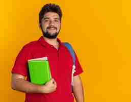 Foto grátis rapaz barbudo estudante com uma camisa pólo vermelha com mochila segurando livros sorrindo alegremente feliz e confiante em pé sobre a parede laranja