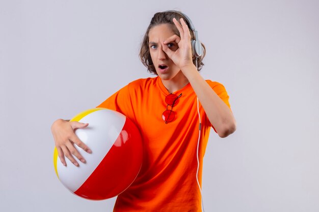 Rapaz bonito com uma camiseta laranja segurando uma bola inflável com fones de ouvido fazendo sinal de ok olhando através deste sinal surpreso em pé sobre um fundo branco