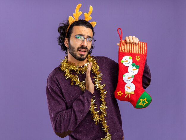 Rapaz bonito descontente com uma argola de cabelo de Natal com uma guirlanda no pescoço, segurando as meias de Natal com a mão estendida para as meias isoladas no fundo azul