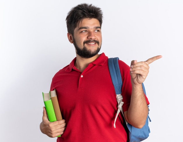 Rapaz jovem barbudo feliz e positivo com uma camisa pólo vermelha com mochila segurando notebooks olhando para o lado apontando com o dedo indicador para o lado em pé