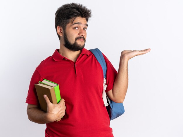 Rapaz jovem barbudo feliz e positivo com uma camisa pólo vermelha com uma mochila segurando notebooks, apresentando o espaço da cópia com o braço da mão em pé sobre a parede branca