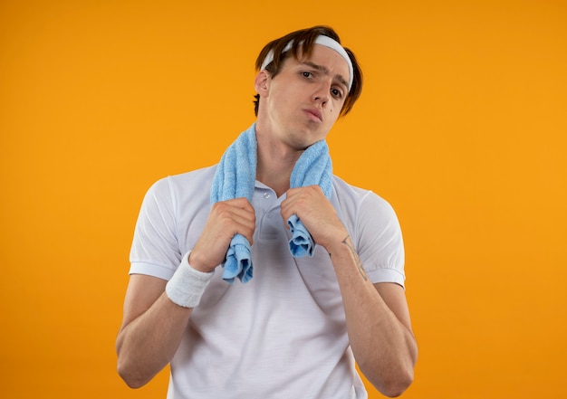 Rapaz jovem e desportivo descontente com fita para a cabeça e pulseira com toalha no ombro, isolada na parede laranja