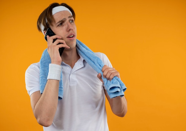 Rapaz jovem e desportivo descontente, olhando para o lado, usando bandana e pulseira com toalha no ombro, isolada na parede laranja com espaço de cópia
