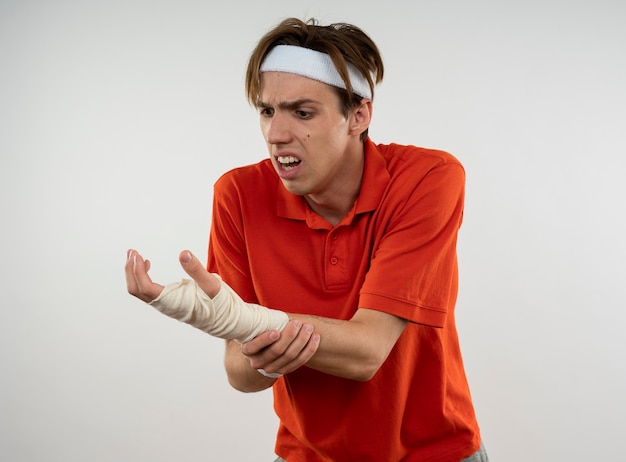 Rapaz jovem e desportivo ferido com fita para a cabeça com pulseira e pulso envolto por bandagem para agarrar o pulso isolado na parede branca