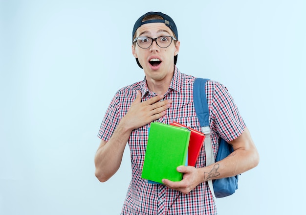 Rapaz jovem estudante surpreso usando bolsa, óculos e boné, segurando livros e colocando a mão no peito em branco