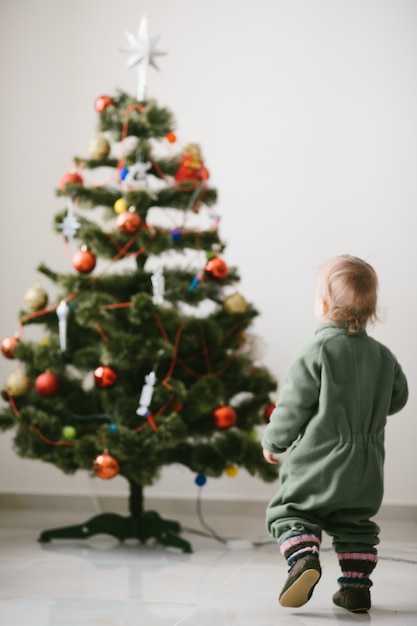 Rapaz pequeno em saltadores verdes anda até a árvore de Natal