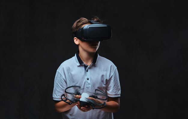 Rapaz vestido com uma camiseta branca usando óculos de realidade virtual e segura um quadricóptero. Isolado em um fundo escuro.