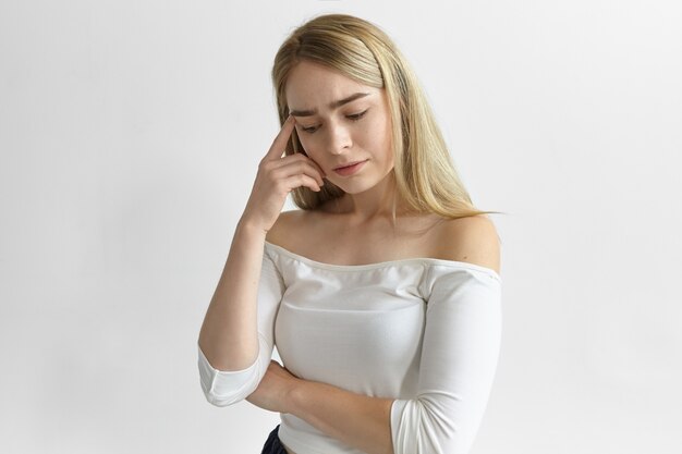 Reação humana, emoções e sentimentos. Foto de uma jovem mulher caucasiana atraente, vestindo uma blusa elegante, frustrada com problemas financeiros, segurando o dedo na têmpora, olhando pensativo