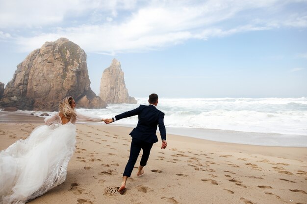 Recém-casados felizes segurando pelas mãos estão correndo pela praia no Oceano Atlântico