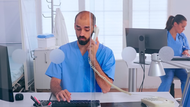 Recepcionistas médicas trabalhando na clínica do hospital, respondendo ao telefone, escrevendo no computador, marcando consultas. Médico uniformizado, escrevendo lista de pacientes consultados e diagnosticados, fazendo pesquisas.