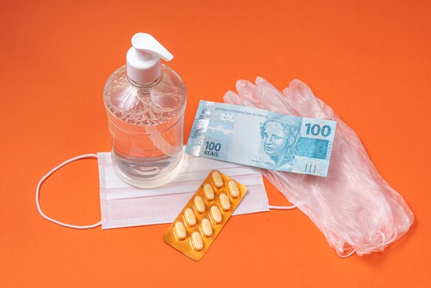 Recipiente de gel de álcool, máscara cirúrgica, remédio e dinheiro real brasileiro, na parede laranja