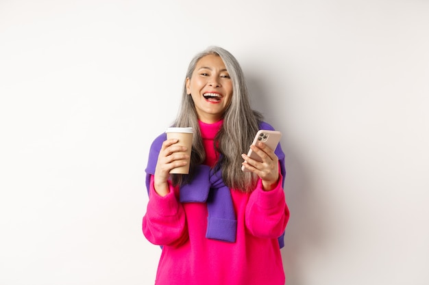 Rede social. Mulher asiática sênior feliz bebendo café e segurando o smartphone, rindo da câmera, em pé sobre um fundo branco