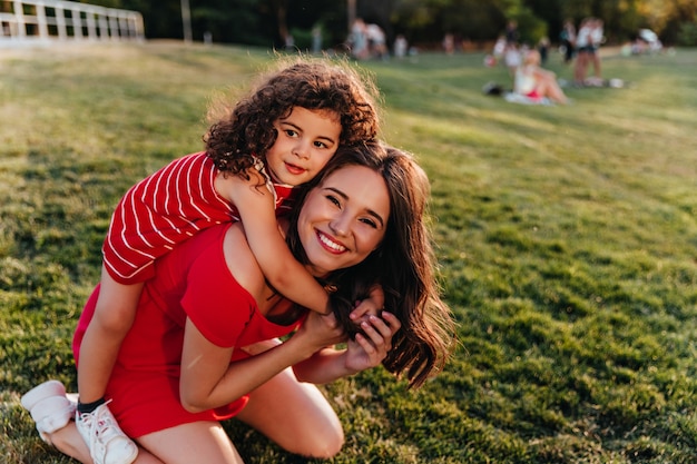 Foto grátis refinada menina abraçando a irmã na natureza feliz modelo feminino com cabelo castanho, brincando com uma criança encaracolada no parque.
