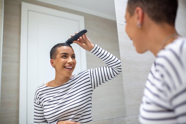 Reflexo de espelho de mulher de cabelos curtos em regata usando máquina de corte de cabelo