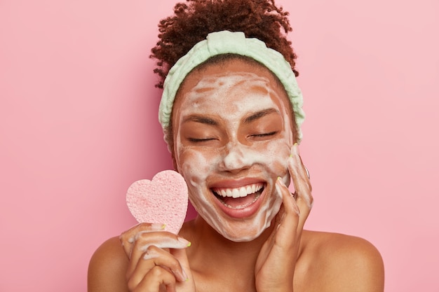 Foto grátis relaxada e alegre modelo feminina de pele escura lava o rosto com bolhas de sabão, desfruta de sessões de mimos, mantém os olhos fechados de prazer, segura uma esponja cosmética, se preocupa com o corpo, fica nua dentro de casa