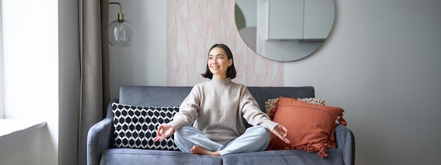 Foto grátis relaxamento e paciência sorrindo jovem asiática em quarto aconchegante sentada no sofá e meditando fazendo