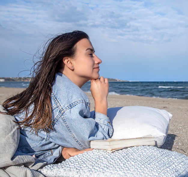 Foto grátis relaxe o conceito, uma mulher na praia em um clima ventoso, descansando sobre um travesseiro com um livro.