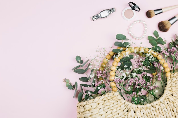 Relógio de pulso; pincel de maquiagem; pulseira de pérolas; pó facial compacto com limonium e gypsophila flores no saco de vime contra um fundo rosa