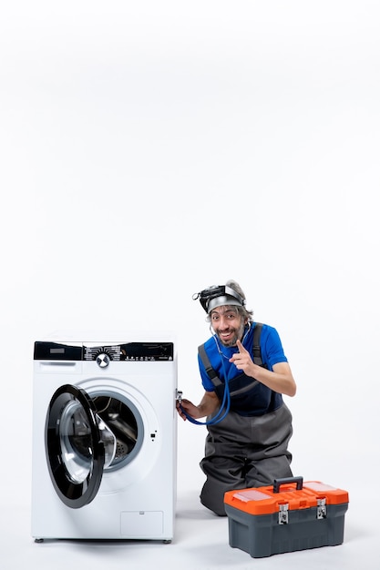 Reparador feliz, vista frontal, colocando o estetoscópio na lavadora, sentado perto da lavadora no espaço em branco