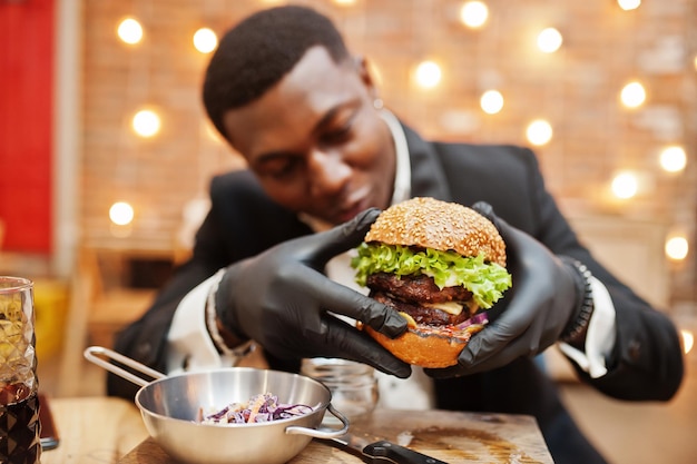 Foto grátis respeitável jovem afro-americano de terno preto e luvas para comida sentado no restaurante com saboroso hambúrguer duplo e refrigerante