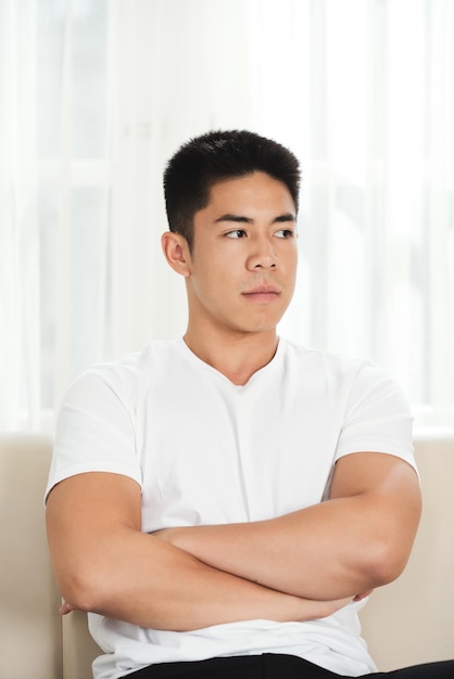 Ressentido jovem asiático sentado no sofá com os braços cruzados