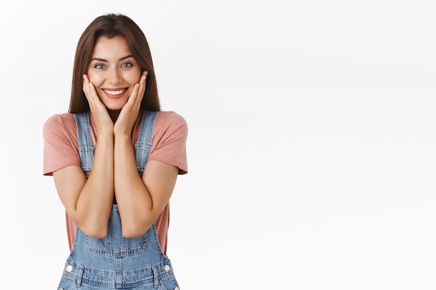 Retratada da cintura para cima feminina, fofa e feliz mulher sorridente tocando a pele pura e limpa e sorrindo deliciada ao se livrar de manchas e acne, sentindo-se revivida e linda, com um fundo branco de pé