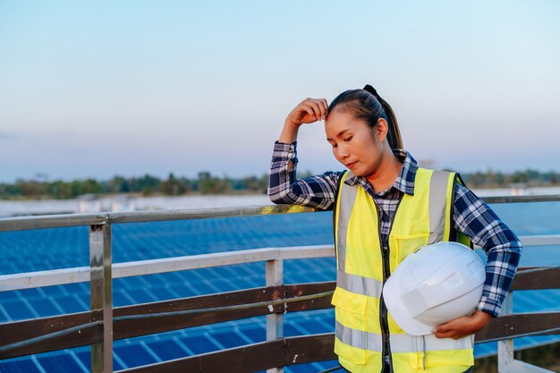 Retrato A jovem engenheira asiática bonita segura o capacete branco na mão, sentindo-se cansada e infeliz após terminar o trabalho na estação do painel solar