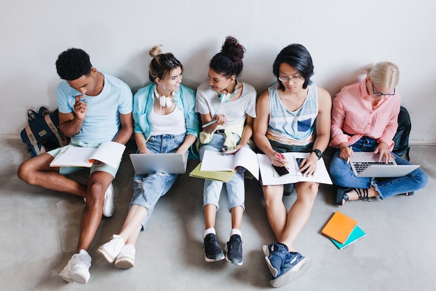 Retrato aéreo de estudantes internacionais à espera de teste na faculdade. Grupo de colegas de universidade sentados no chão com livros e laptops, fazendo lição de casa.
