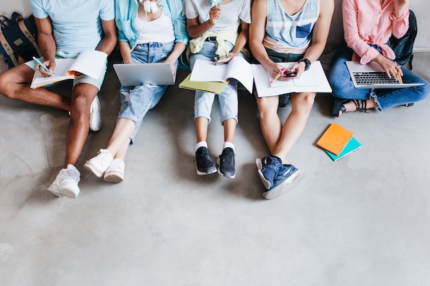 Retrato aéreo de jovens com laptops e smartphones, sentados juntos no chão. Alunos escrevendo palestras segurando livros didáticos sobre os joelhos.