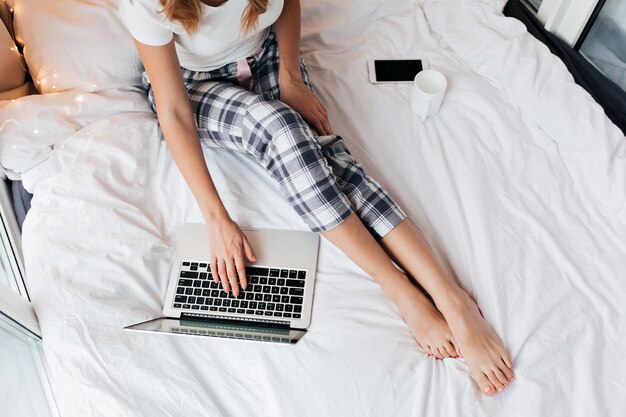 Retrato aéreo de mulher em calças xadrez, usando o laptop. foto interna de freelancer feminino de pijama, trabalhando na cama.