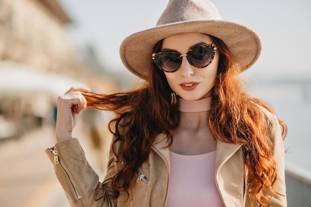 Retrato ao ar livre da adorável mulher de cabelos compridos com expressão facial séria em pé na desfoque de fundo. Foto da elegante garota branca em óculos de sol brinca com seu cabelo ruivo brilhante.