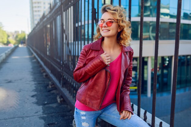 Retrato ao ar livre da mulher entusiasta de hipster alegre na jaqueta de couro na moda. Mostrando sinais à mão.