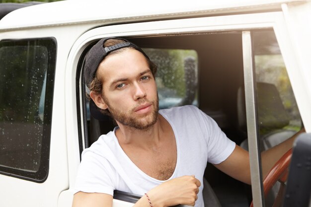 Retrato ao ar livre de atraente jovem hippie com barba elegante olhando com sorriso confiante e orgulhoso enquanto está sentado dentro de seu veículo safari branco