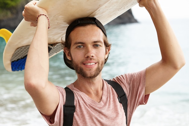 Retrato ao ar livre de belo jovem surfista vestindo snapback para trás posando contra o mar azul, segurando seu bodyboard branco sobre a cabeça, sorrindo alegremente