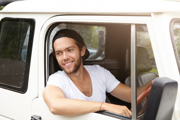 Retrato ao ar livre de bonito jovem barbudo no boné de beisebol saindo da cabeça pela janela aberta do seu carro branco sorrindo