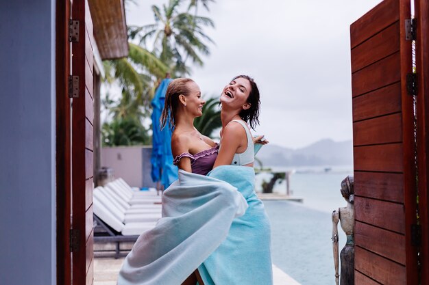 Retrato ao ar livre de duas mulheres felizes em biquínis com toalhas de praia de férias fora da villa perto da piscina em dia chuvoso