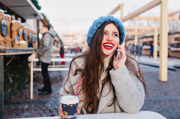 Retrato ao ar livre de excitada menina morena com casaco de lã, aproveitando o fim de semana de inverno em dia quente. Foto de uma senhora caucasiana de cabelos compridos com um lindo chapéu azul, posando na rua borrada