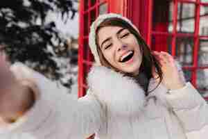 Foto grátis retrato ao ar livre de mulher bonita com sorriso feliz, fazendo selfie em londres durante as férias de inverno. adorável mulher com chapéu branco tirando foto de herslef ao lado da cabine telefônica vermelha.