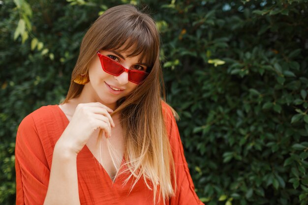 Retrato ao ar livre de mulher bonita no vestido laranja e óculos de sol vermelhos.