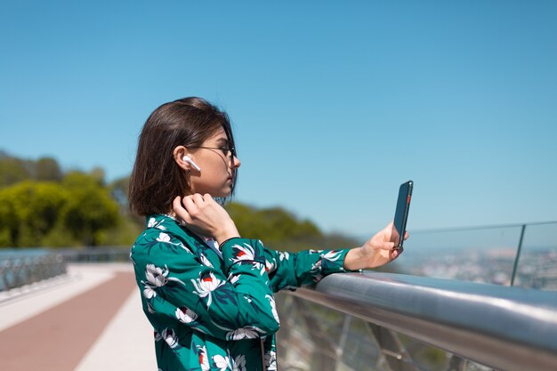 Retrato ao ar livre de mulher com camisa verde casual em dia de sol fica na ponte, olhando na tela do telefone, fones de ouvido bluetooth sem fio