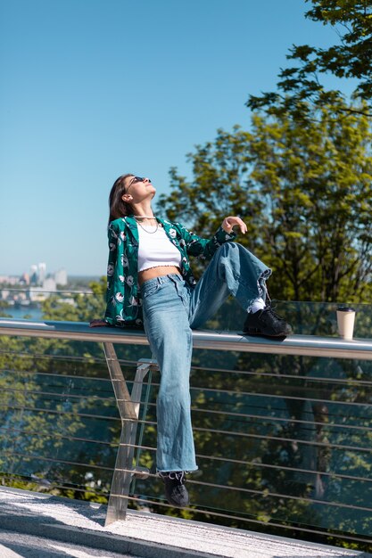 Retrato ao ar livre de mulher em jeans e camisa verde casual em dia de sol na ponte com uma vista incrível da cidade pela manhã