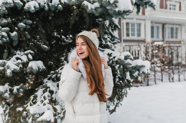 Retrato ao ar livre de mulher engraçada com chapéu de malha, olhando para longe enquanto posava perto de abeto vermelho verde coberto de neve.