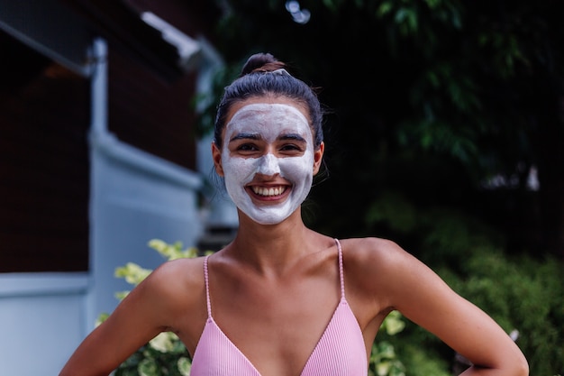 Retrato ao ar livre de pele bronzeada acalma mulher bonita caucasiana de biquíni no spa com máscara esfoliante branca no rosto