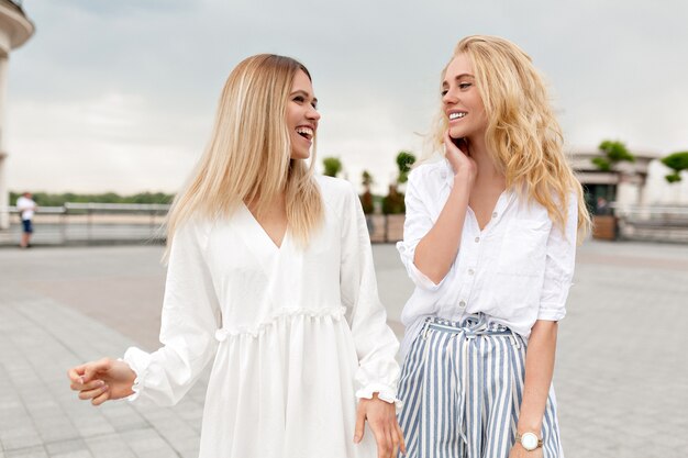 Retrato ao ar livre de senhoras bonitos se divertindo lá fora relaxando na cidade de verão, enquanto caminhava na rua e ria num dia nublado de verão. Duas garotas estilosas lá fora