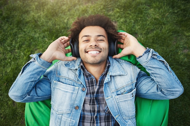 Retrato ao ar livre de um homem alegre e otimista de pele escura, com cabelo eriçado e cabelo afro, deitado em uma cadeira de pufe ou na grama, sorrindo enquanto ouve música com fones de ouvido e os segura com as mãos