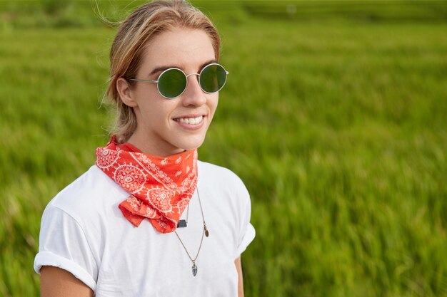 Retrato ao ar livre de uma bela mulher sorridente, vestida com uma camiseta casual e usando óculos escuros, encostada na grama verde, passeios