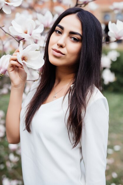 Retrato ao ar livre de uma jovem mulher bonita perto de magnólia com flores.