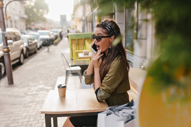 Retrato ao ar livre de uma menina morena chocada em óculos de sol, falando no telefone na manhã de outono. Mulher shopaholic surpresa com pele bronzeada, posando no café, depois de fazer compras no fim de semana.
